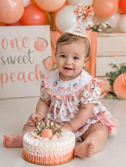 May include: A baby wearing a peach-themed outfit, bib, and party hat, sitting in front of a cake. one sweet peach girls first birthday bubble romper outfit peachy blush pink bows first birthday girl romper peach and white outfit, peach smocked dress first birthday embroidered peaches smocked short sleeve 1st birthday outfit ruffle bubble romper baby girls fruit cake smash outfit, the mod mango baby boutique co