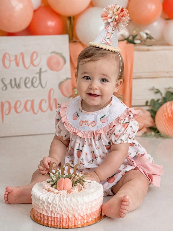 May include: A baby wearing a peach-themed outfit, bib, and party hat, sitting in front of a cake. one sweet peach girls first birthday bubble romper outfit peachy blush pink bows first birthday girl romper peach and white outfit, peach smocked dress first birthday embroidered peaches smocked short sleeve 1st birthday outfit ruffle bubble romper baby girls fruit cake smash outfit, the mod mango baby boutique co