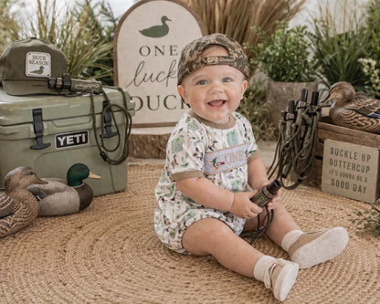 May include: A smiling baby wearing a camouflage hat and a romper with a duck-themed print. The baby is holding a hunting accessory. A cooler, hat, and sign with the words "ONE luck DUCK" are in the background.