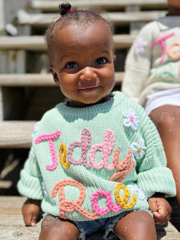 May include: A mint green knit sweater featuring the name Teddy Rae embroidered in chunky, multi-colored yarn letters in pink, peach, and yellow. The sweater is decorated with small, white, five-petaled flowers with yellow centers.